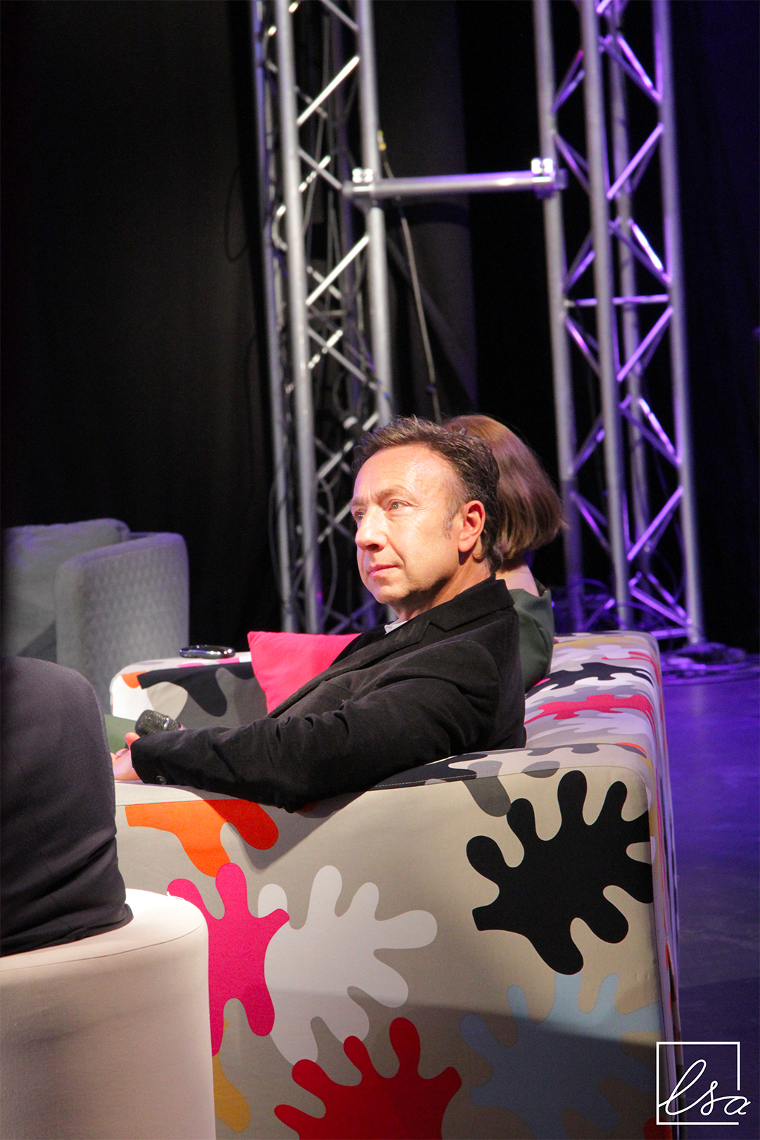 Photographie de Stéphane Bern, animateur radio et télévision, sur scène, assis sur un canapé.