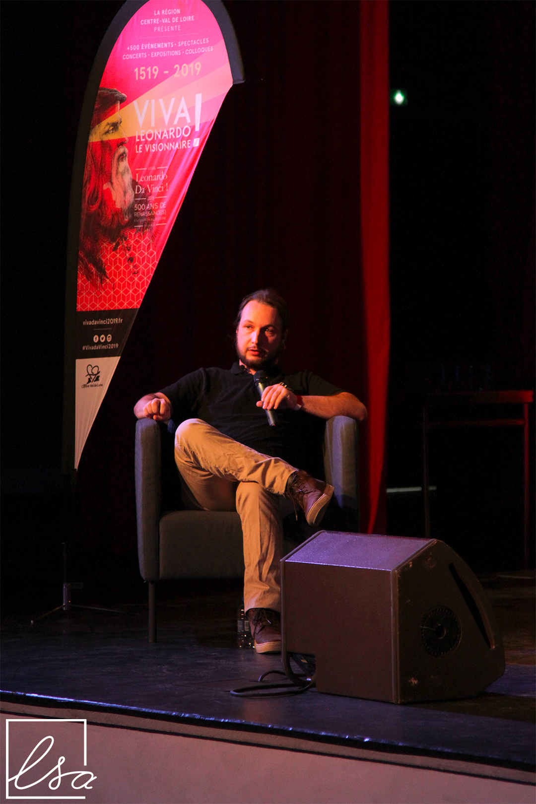 Photographie de Renaud Berthier, Président de ls-a, assis sur scène le micro à la main, en train de donner sa conférence sur le génie logiciel.